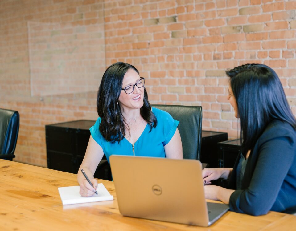 Two women engaged in cross-cultural negotiation strategies, attentively sitting at a conference table with laptops.