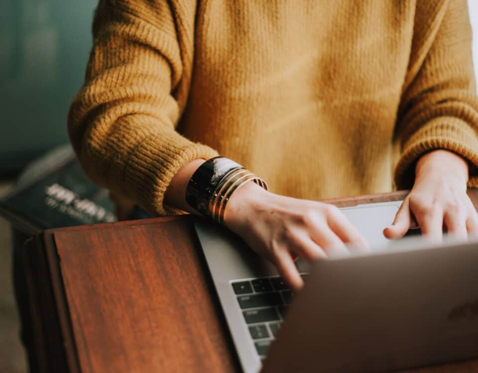 A woman in a yellow sweater immersed in cultural nuances while typing on a laptop during business negotiations.