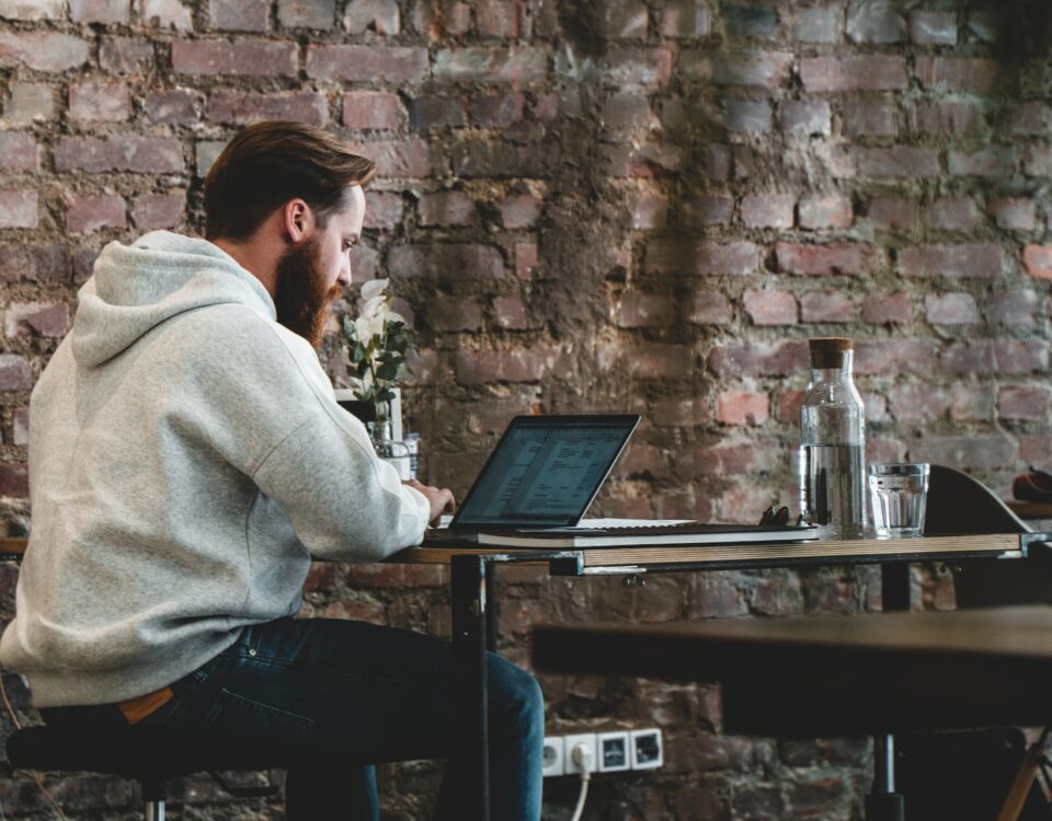 A man engaged in remote work, sitting at a desk with a laptop in front of a brick wall.