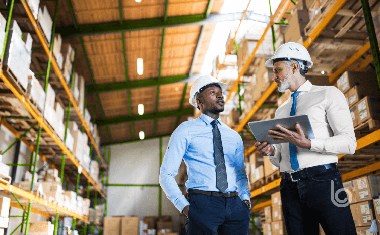 Two men in hard hats discussing global supply chain challenges in a warehouse.
