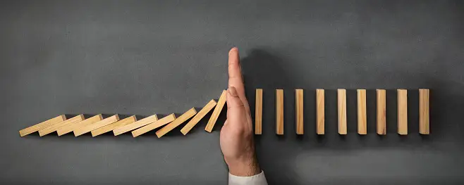 A man's hand is holding a wooden domino, illustrating the effects of trade barriers.