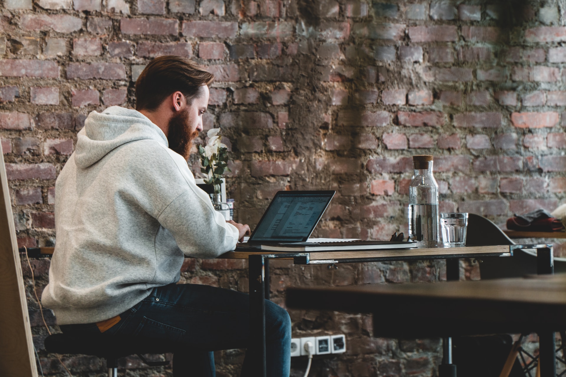 A man engaged in remote work, sitting at a desk with a laptop in front of a brick wall.