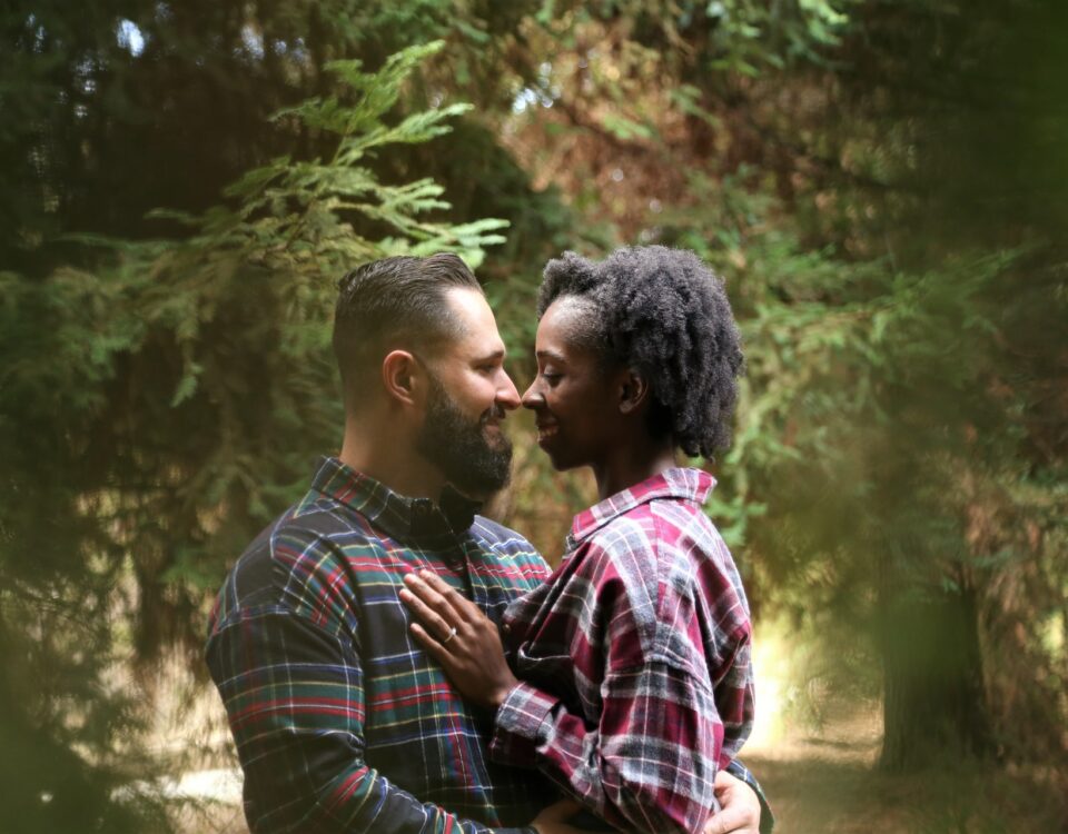 A diverse couple embraces in the woods during their engagement session, showcasing the inclusion and diversity that can be seen in international trade.
